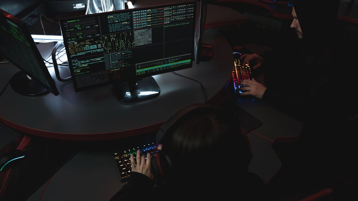 Two people at glowing keyboards in a dark room, one watching streams of data on a monitor